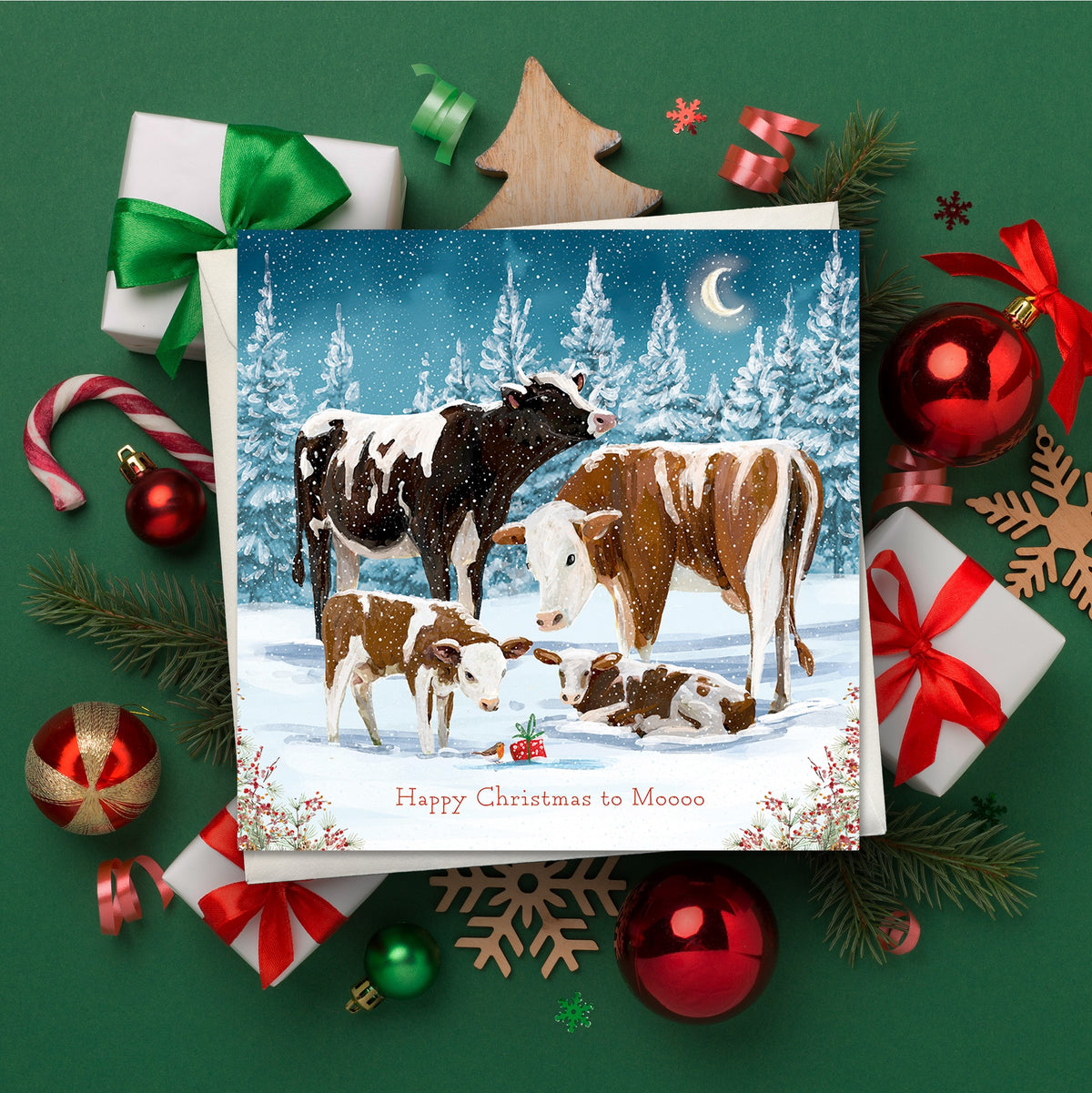 Christmas card featuring a cow family standing in a snowy winter landscape, surrounded by wrapped presents, baubles, pine branches and festive decorations. Text reads: “Happy Christmas to Moooo”.