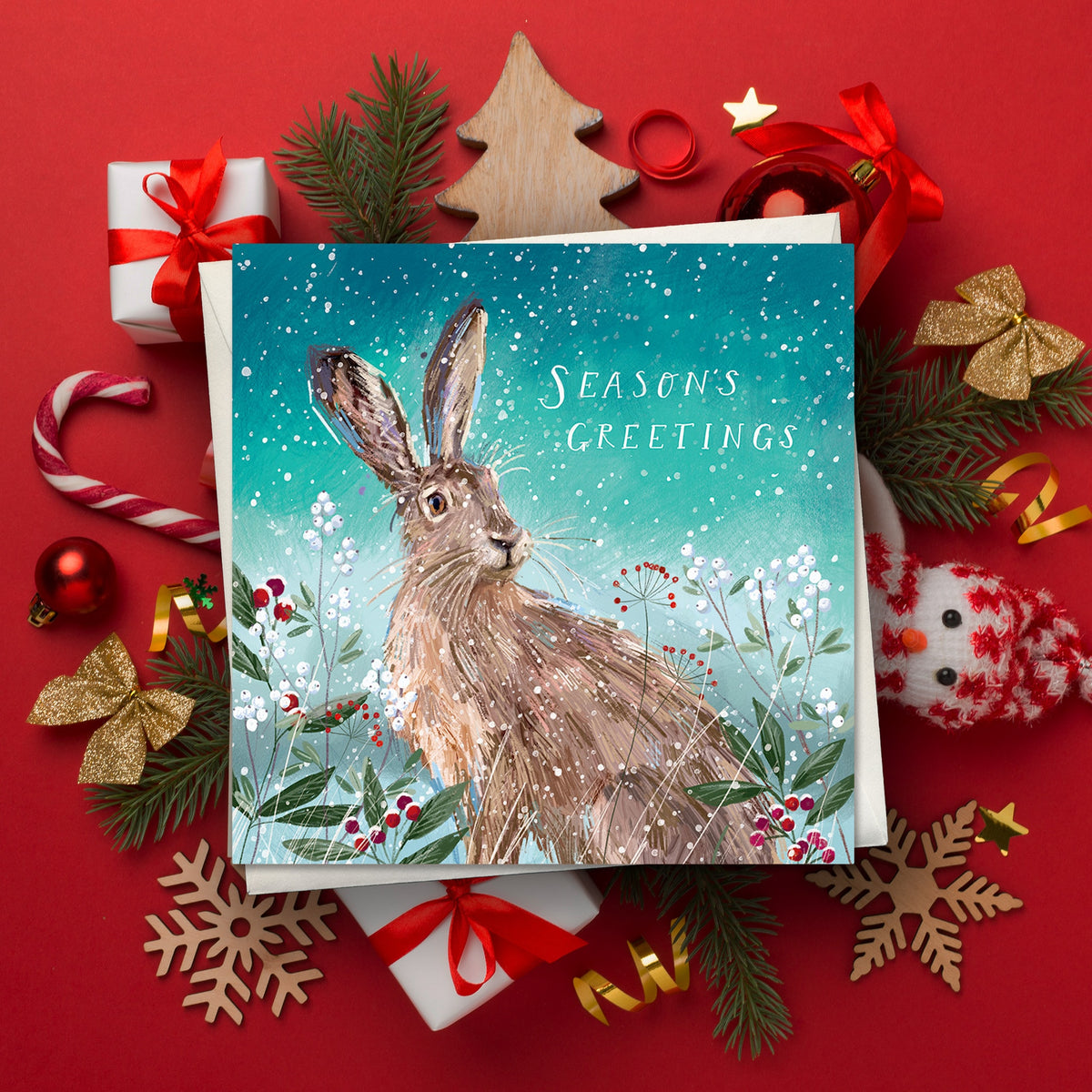 Christmas card featuring an illustrated brown hare sitting among winter berries and frosted plants during falling snow. The card reads "Season's Greetings" and is surrounded by wrapped presents, pine branches, baubles, a candy cane and festive decorations on a red background.